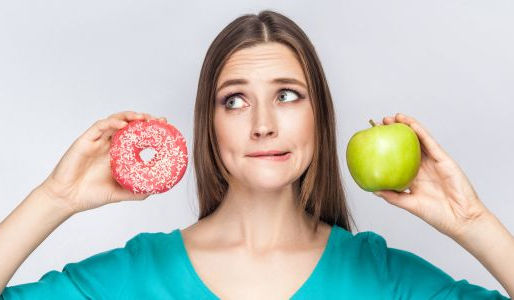 A young woman in a teal shirt holds a pink-frosted donut in one hand and a green apple in the other, looking conflicted and biting her lip. Her facial expression reflects indecision, highlighting the struggle between choosing ultra-processed foods and healthier options. The background is a neutral grey, keeping the focus on her expression and the food choices.