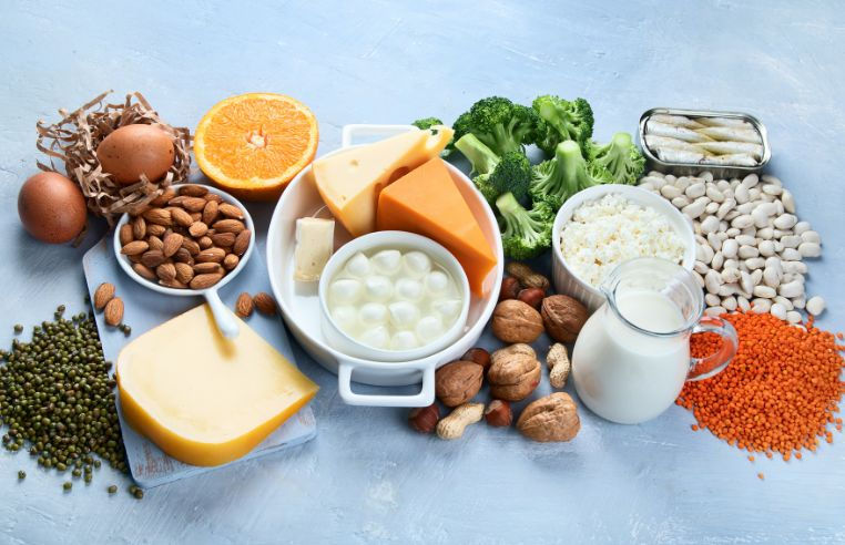 Flat lay of calcium-rich foods including eggs, almonds, cheese, broccoli, sardines, beans, lentils and milk on a light blue background