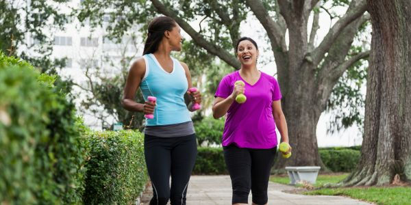 Two women in their 40s walking outdoors carrying dumbbells, green park setting