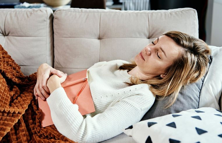 A woman rests on a light grey sofa covered with a rust-coloured blanket, her hand resting gently on her abdomen, eyes closed. Calm, domestic recovery setting.