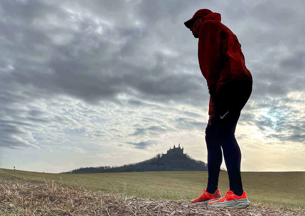Läufer steht ruhig im Gelände, eine Hand am Knie, Blick leicht nach unten – nicht schmerzgeplagt, sondern nachdenklich. Fokus auf Achtsamkeit und Körperwahrnehmung. Bild soll vermitteln: Schmerz ist ein Signal, kein Feind.