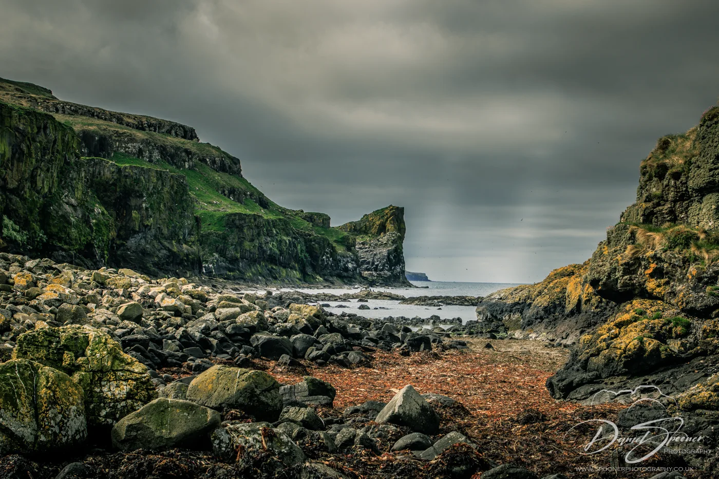 Rocky cliffs and coastal terrain on Lunga Island beneath a stormy sky, with green moss and seaweed covering the foreground.