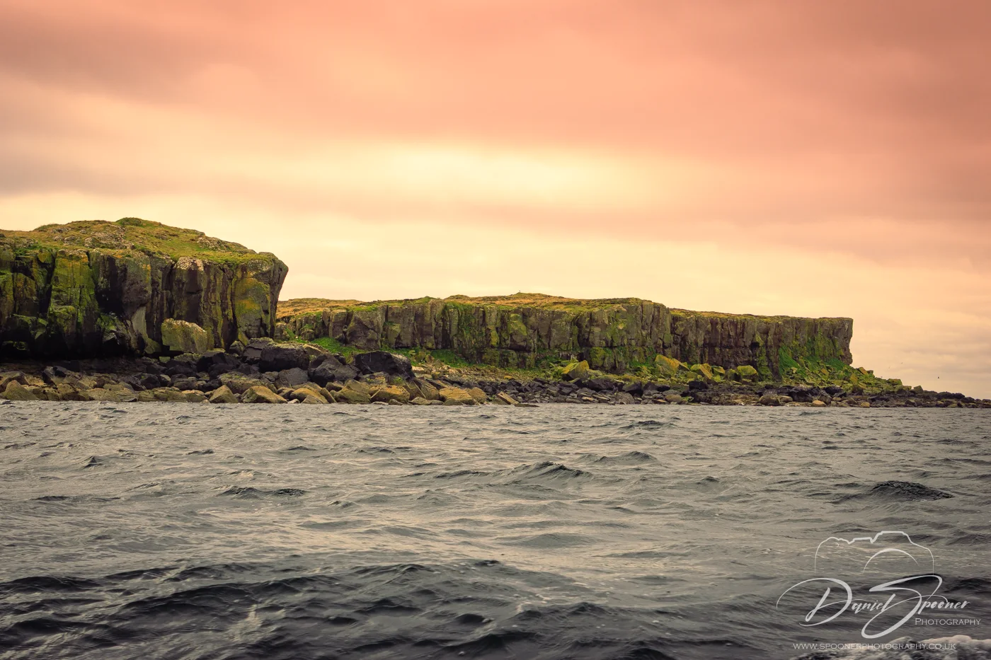 Dramatic green cliffs of Staffa island under a warm-toned sunset sky, rising from the dark sea in the Inner Hebrides.
