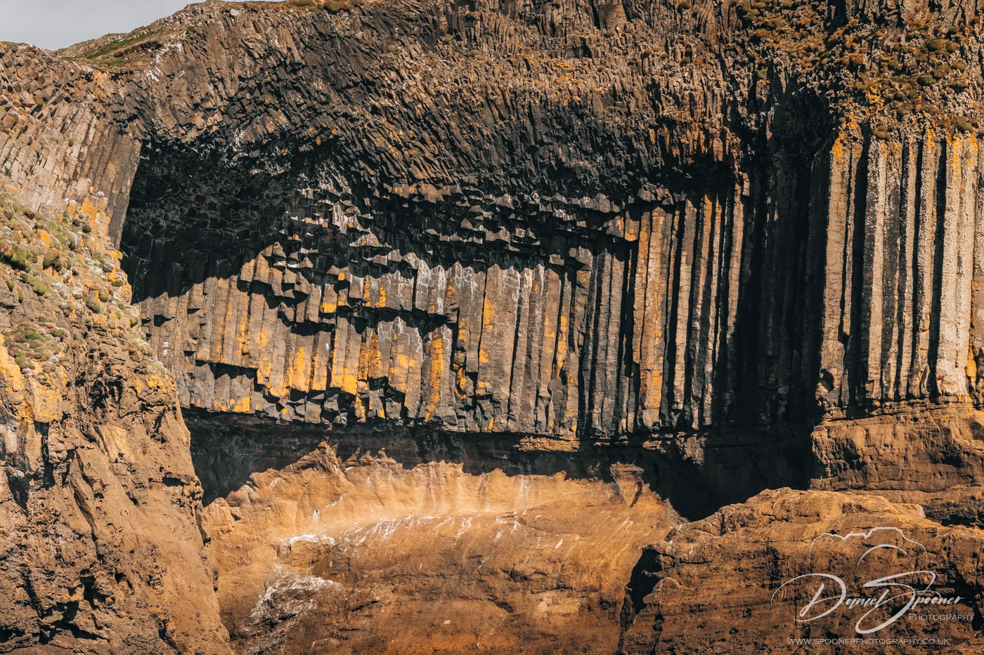 Vertical basalt columns on Staffa partially covered in orange lichen, with overhangs and shadows emphasizing their dramatic formation.