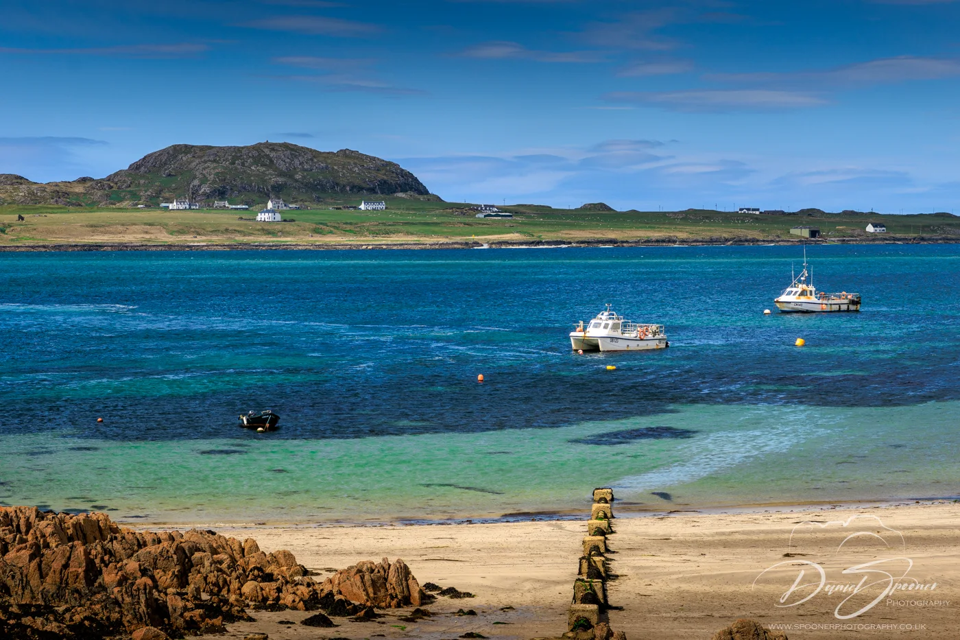 View across vibrant turquoise sea toward the island of Iona, showing white houses nestled at the base of a rocky hill and several fishing boats floating on the water under a bright blue sky.