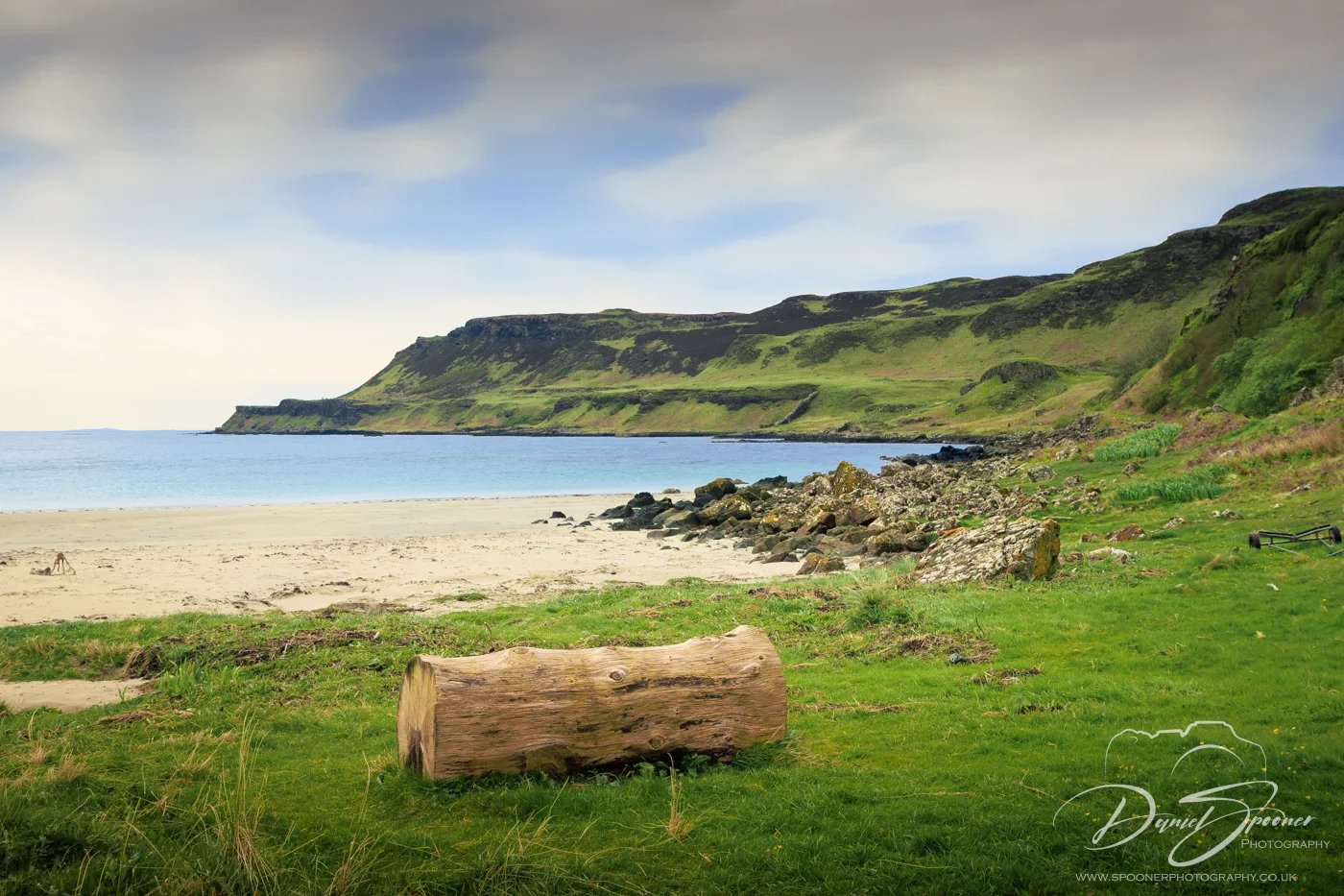 View of Calgary Bay on the Isle of Mull with a log resting on grass in the foreground, a sandy beach, and green cliffs meeting the sea under a calm sky.
