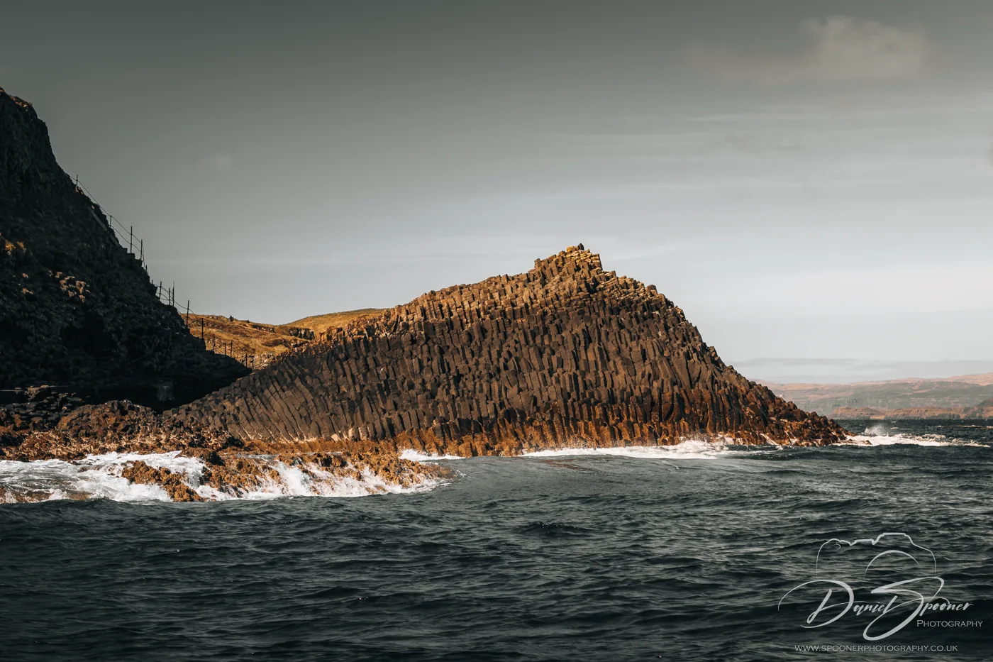 Angular basalt columns rising from the sea at Staffa under a muted sky, with waves crashing along the dark volcanic rock.