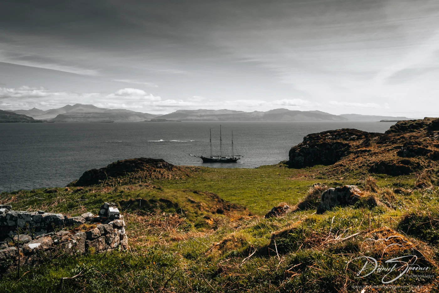 A ship viewed from a grassy cliffside on Lunga Island with surrounding stone walls, overlooking the sea and distant mountains.