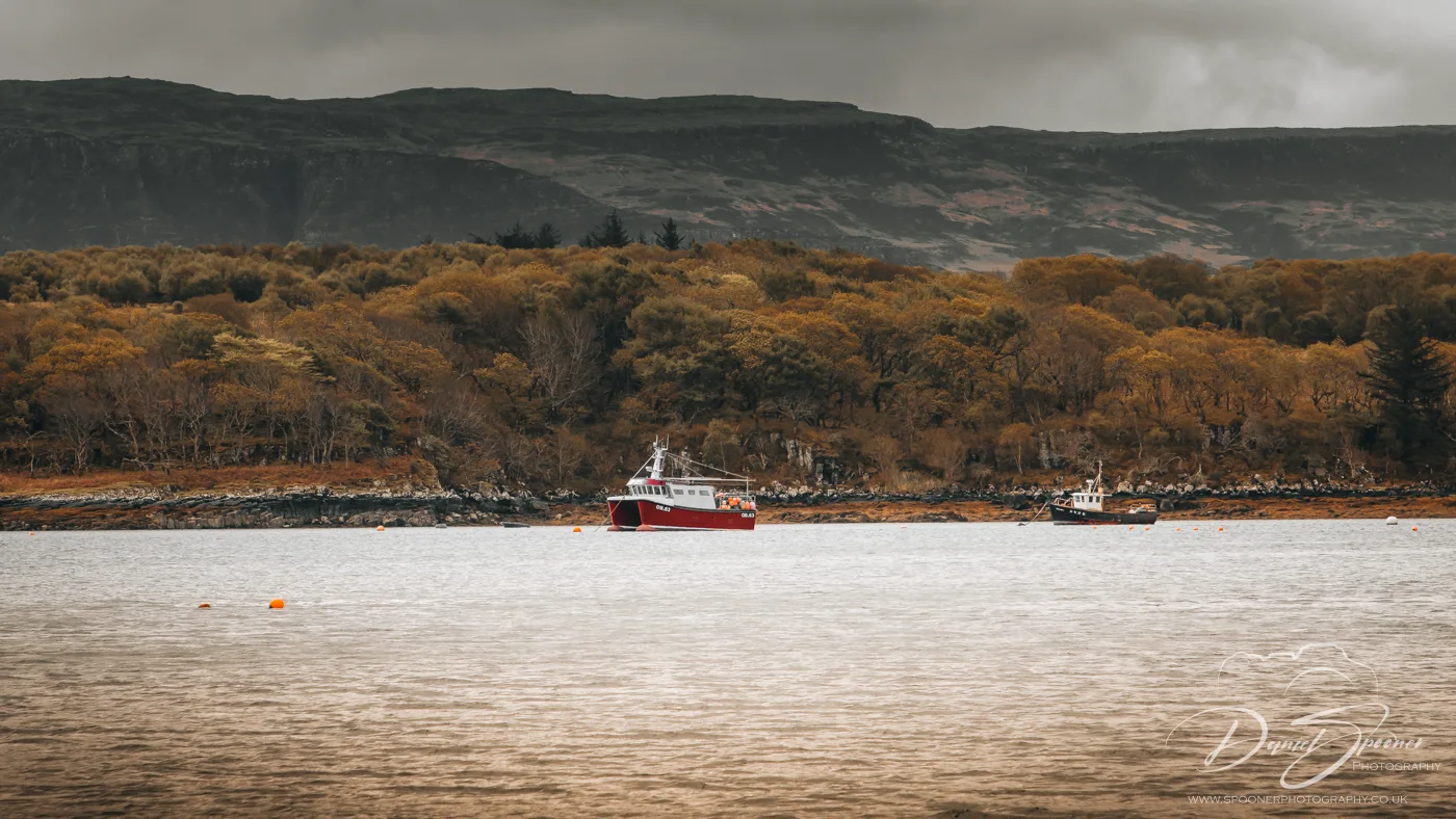A red and white fishing boat floats on a calm loch with autumnal trees and a dark mountainous backdrop on the Isle of Mull.