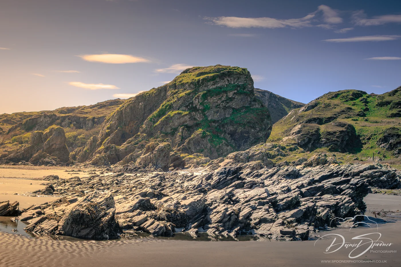 The rugged rocky formations and sandy beach of Uisken under warm sunlight, with grassy cliffs rising in the background and a calm sky overhead.