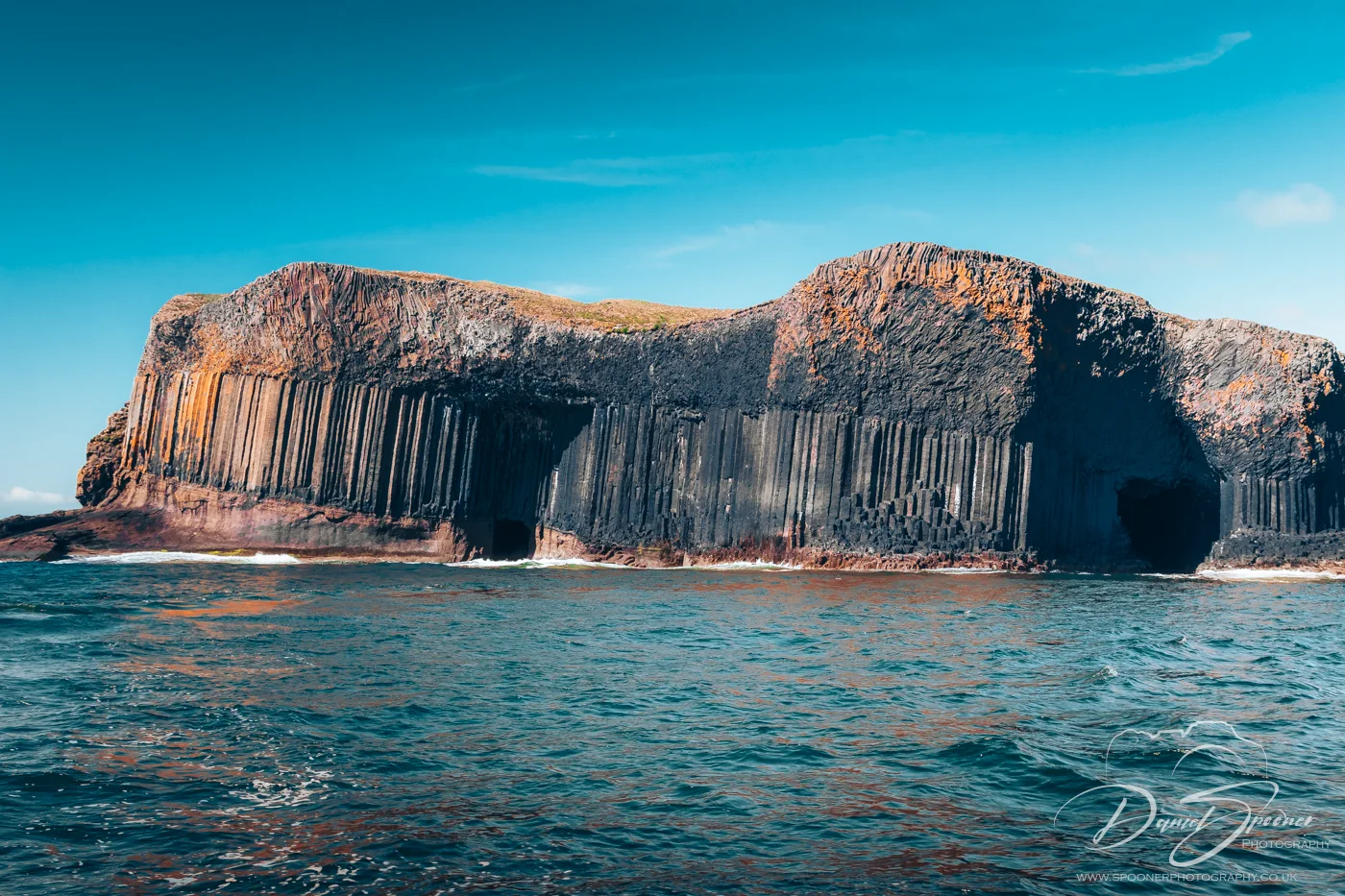 View of Staffa island showing Fingal's Cave and vertical basalt columns set against bright blue skies and turquoise waters.