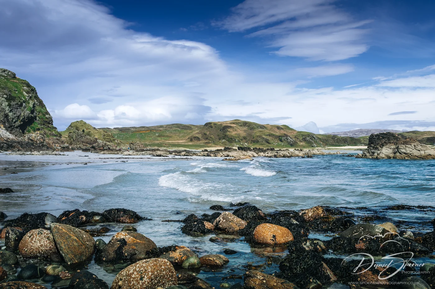 A scenic view of Uisken Beach showing rocky shoreline with seaweed-covered boulders, gentle waves lapping in, and grassy hills in the background beneath a vivid sky.