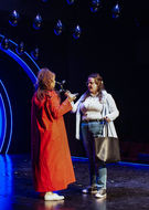 Sara Jane Moore (Siobhan McNulty in jeans, a white turtleneck and blue vest. Brown hair) threatening Squeaky Fromme (Gabriela Sorrentino, in a red robe, dark blonde hair) with a black balloon animal representing the dog Sara Jane recently killed