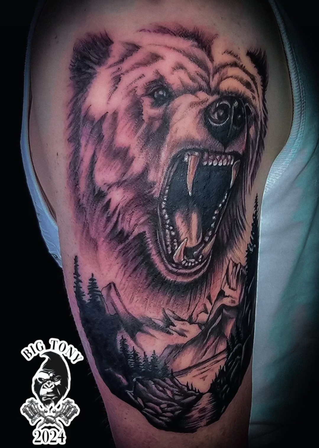 Black-and-grey realistic roaring bear over a mountain and forest landscape tattooed on an upper arm, carrying the artist's own Big Tony 2024 mark