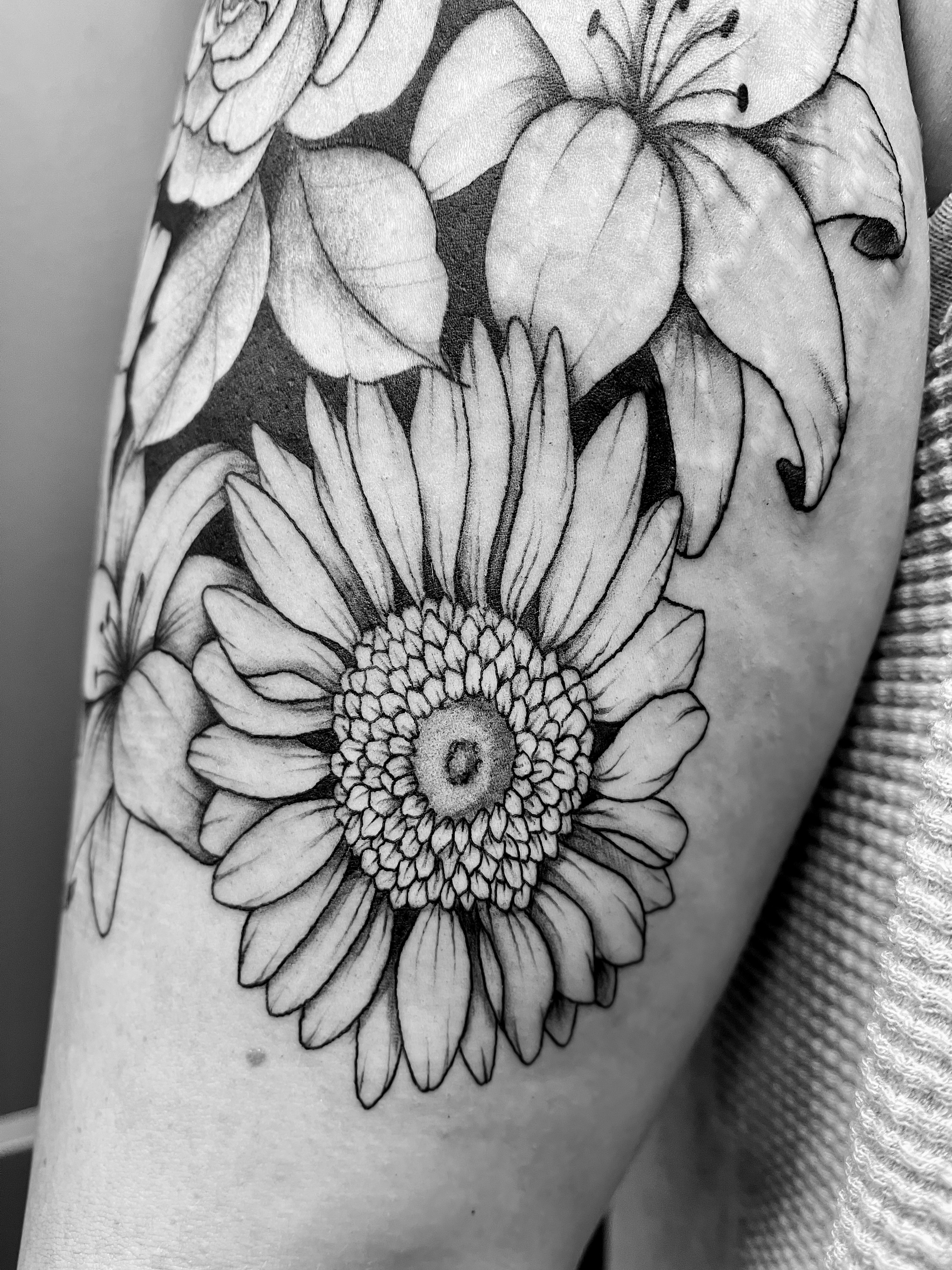 Black-and-grey floral tattoo of a sunflower and lilies on an upper arm