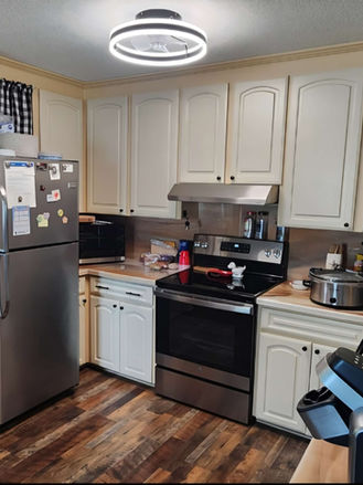 Before kitchen remodel showing original cream cabinets, laminate countertops, stainless steel appliances, patterned wallpaper, and dark wood-look flooring prior to renovation.