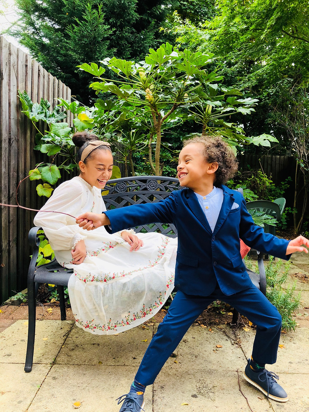 Girl wearing a white embroidered dress sitting on a black bench laughing while a boy in blue suit with blue dress shoes is standing in front of her with a stick. The boy is also laughing as he strikes a pose