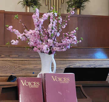 Two Voices United books in front of a white vase with pink blossoms in it on a wooden piano top