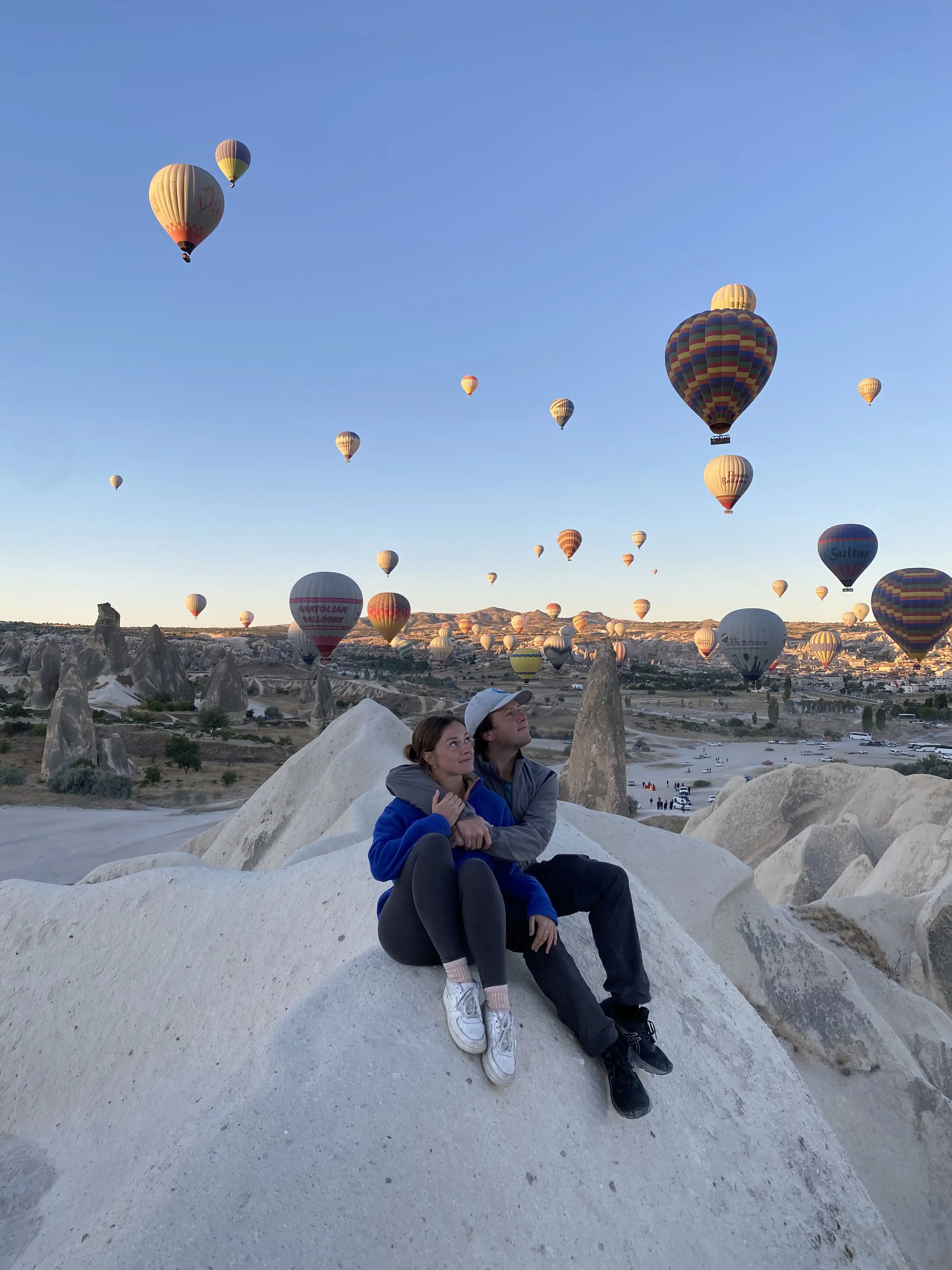 Couple sitting on a rock with many hot air balloons behind them in Cappadocia, Turkey