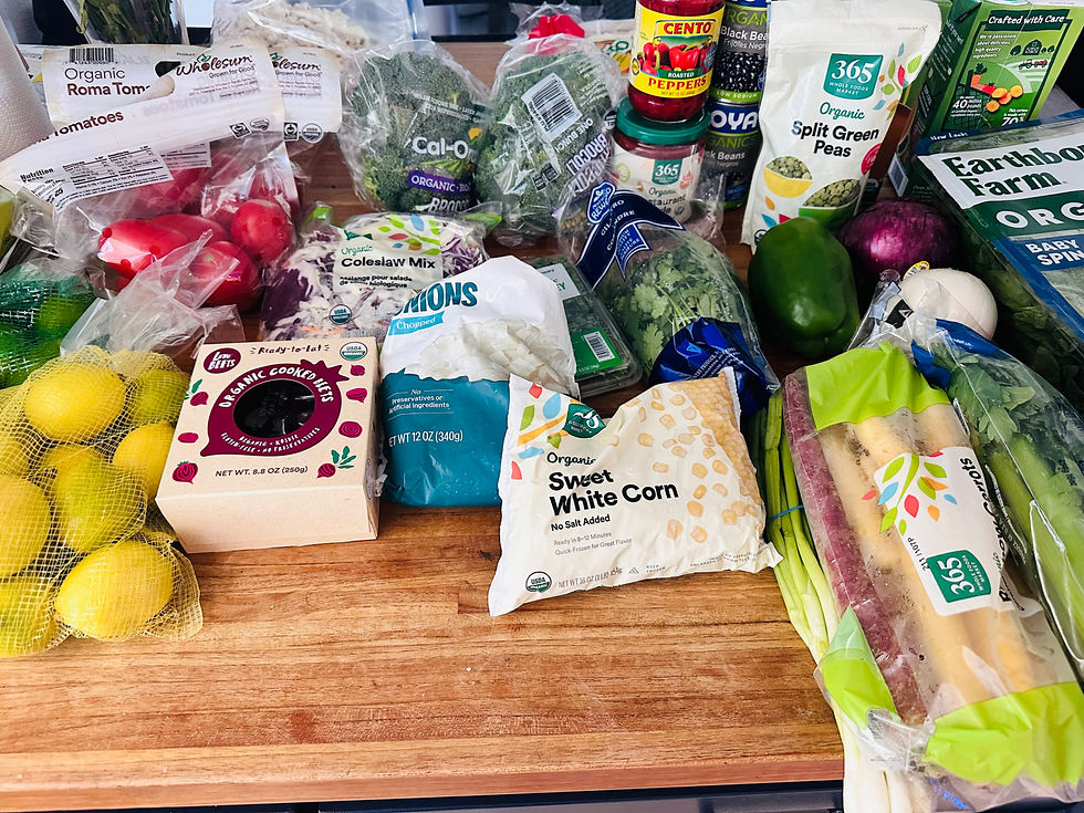 Assorted organic groceries on a wooden counter, including tomatoes, greens, beans, corn, lemons, and labeled bags and jars.