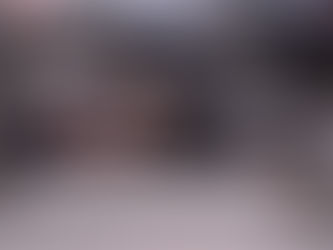 El hombre está tumbado de lado sobre el cemento o pavimento. Parece joven, de complexión delgada, y está durmiendo profundamente con los ojos cerrados. Viste pantalones oscuros y zapatillas deportivas negras con detalles blancos. Su torso está descubierto, revelando su abdomen. Tiene lo que parece ser un vendaje blanco en el antebrazo derecho. Utiliza una prenda de ropa o un trozo de tela oscuro como almohada.