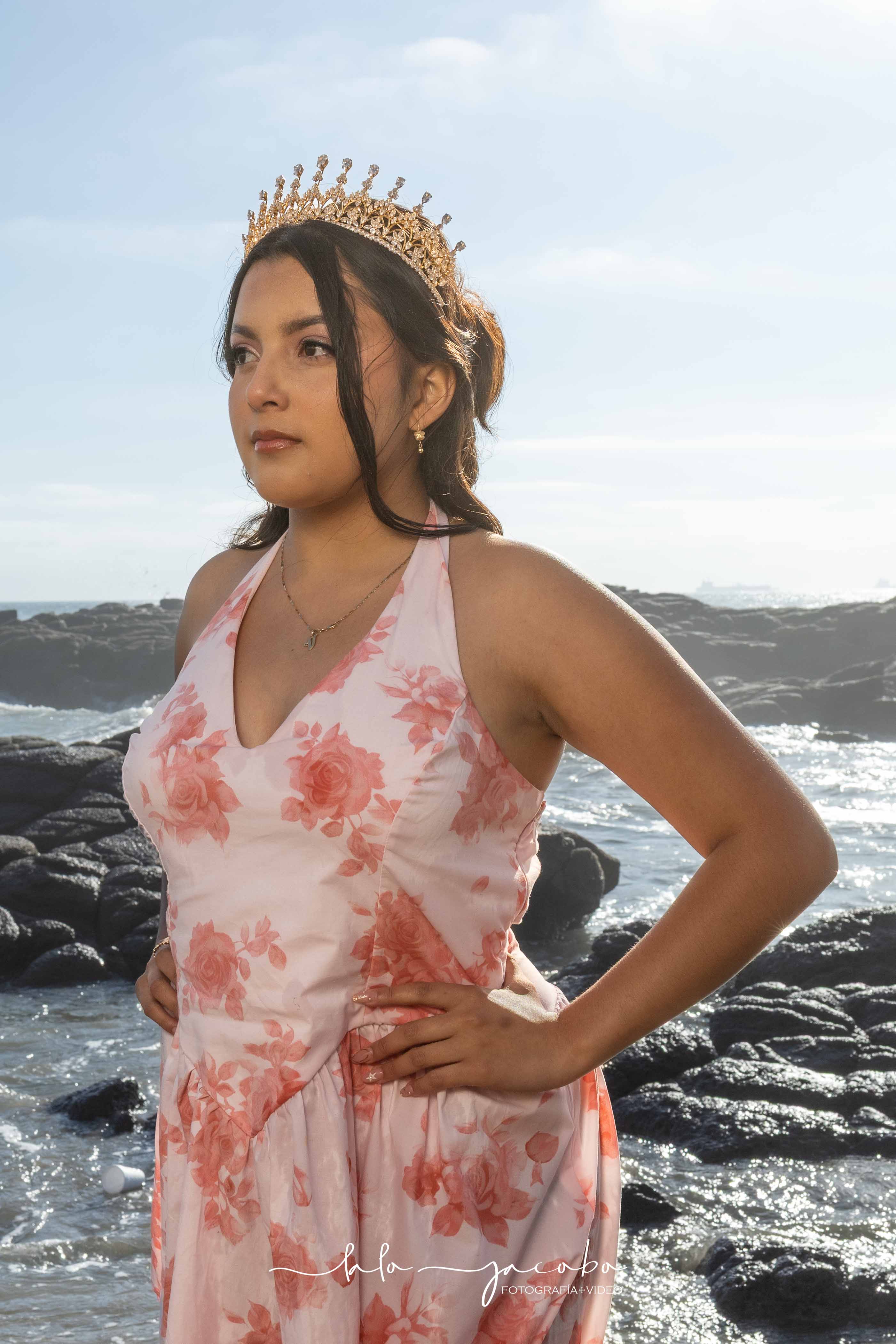 Retrato de quinceañera con corona dorada y vestido floral rosa junto al mar, por Lalo Jacobo Fotografía + Video.