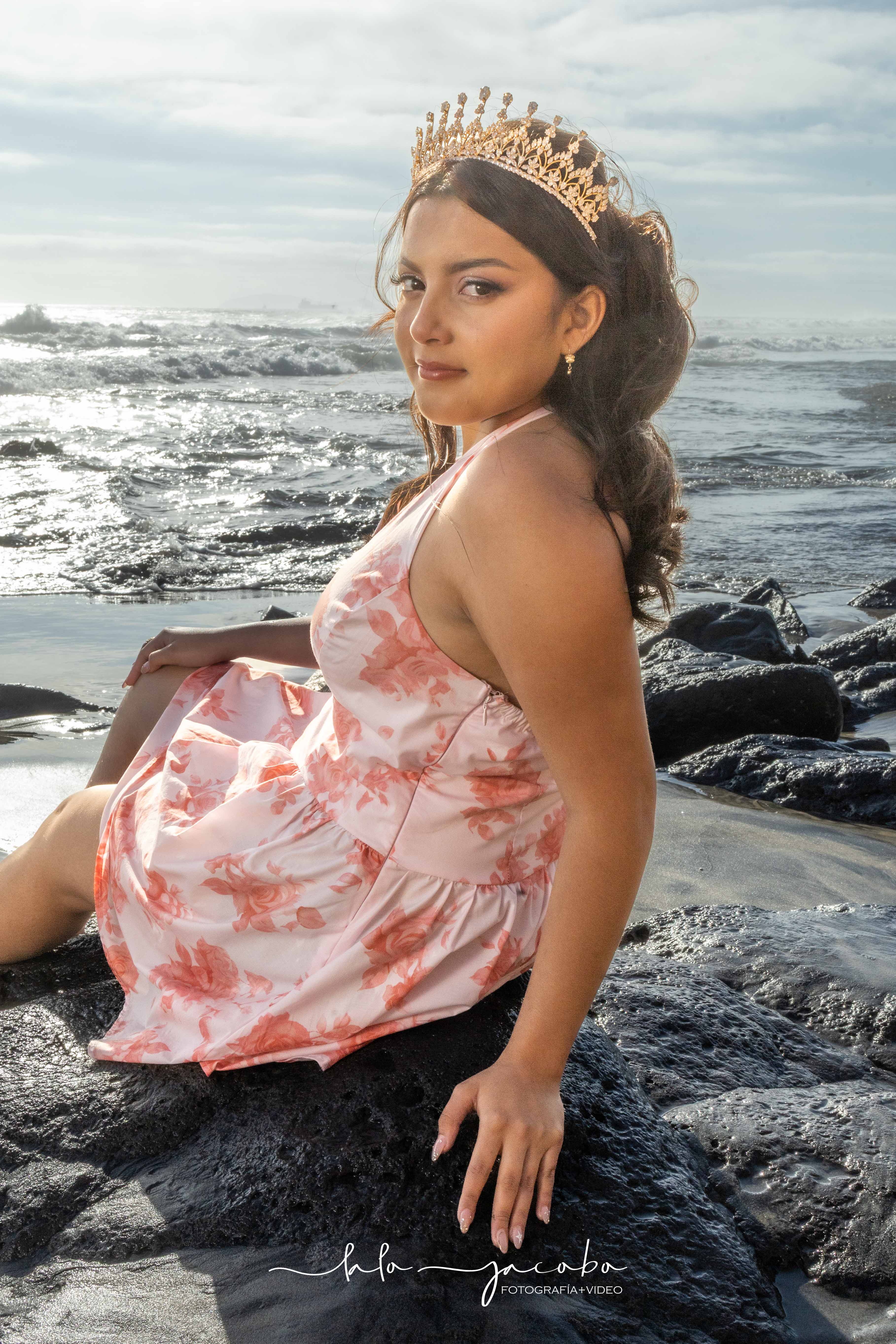 Quinceañera sentada junto al mar con vestido floral y corona, fotografiada por Lalo Jacobo Fotografía + Video.