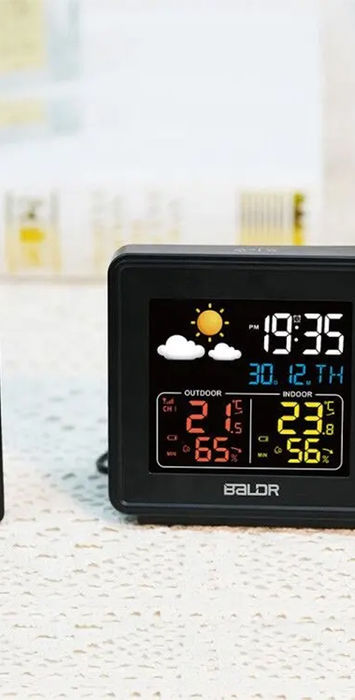 Digital wireless weather station with colour LCD screen displaying indoor and outdoor temperature and humidity, weather icons, and historical data. The device includes a separate black sensor unit for remote monitoring.