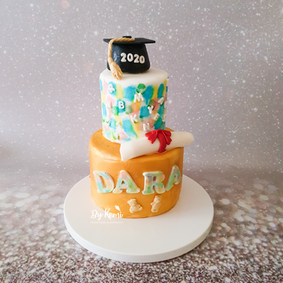 Two tier graduation cake. The bottom tier is gold with the name "Dara" on the bottom. The top tier is white with multi coloured blobs. The cake is topped with a black graduation cap and diploma.