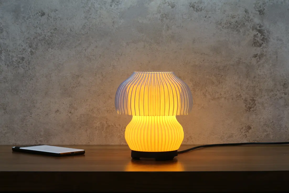 Jellyfish-shaped 3D printed table lamp glowing with warm light, placed on a wooden surface against a textured wall background