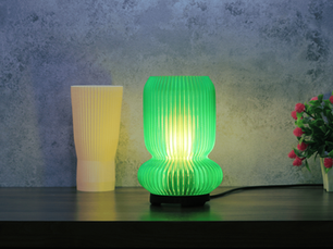 Green lamp glowing on a wooden table next to porcelain skin coloured inner lamp layer and a small plant with pink flowers, set against a textured grey wall.