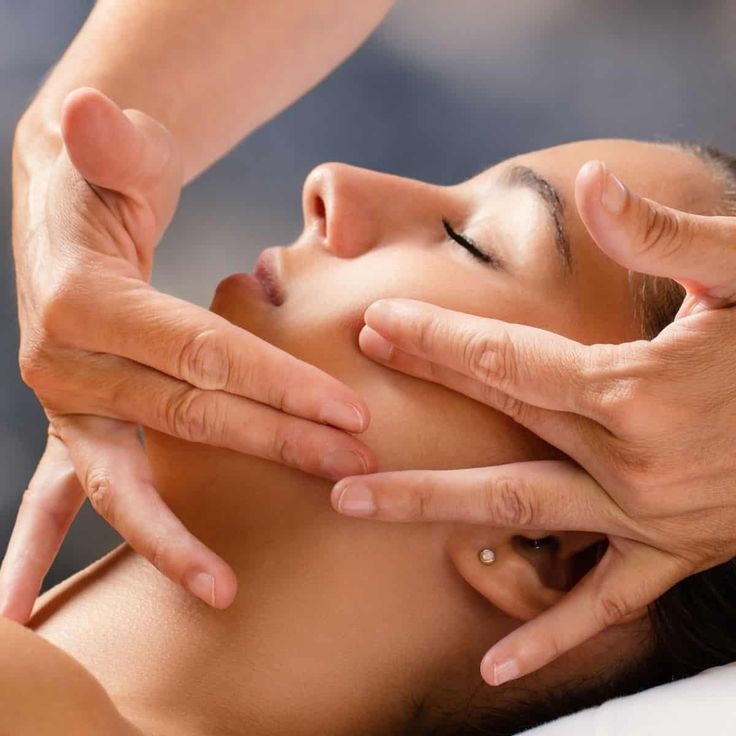 A person receives a relaxing facial massage, eyes closed, calm expression. Hands gently hold the face. Soft, blurred background.