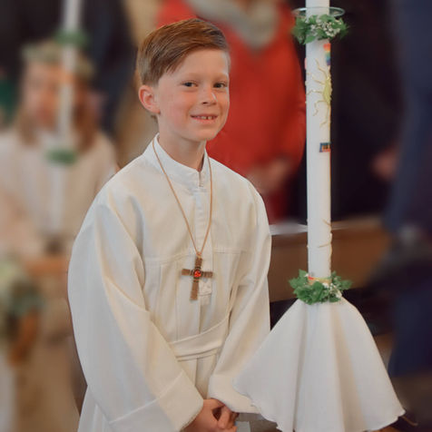 Ein Schuljunge steht im weißen Gewand mit gefalteten Händen im Fokus des Bildes. Um seinen Hals trägt er eine Kette mit einem großen, schweren Kreuz als Anhänger. Er lächelt freundlich in die Kamera. Im Hintergrund zeichnen sich verschwommene Kirchbänke und weitere Kirchbesucher ab.