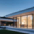 Modern one-story home at dusk with floor-to-ceiling glass walls showing a lit living room, lawn, and minimalist landscaping