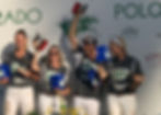 Four smiling people in EXIT shirts hold trophies and blue boxes at a polo event backdrop. Bright, sunny mood with floral decor.