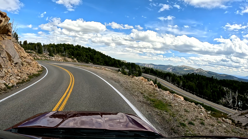 View from a car on a winding mountain road, curving past rocks and pines under a bright blue sky with billowing clouds.
