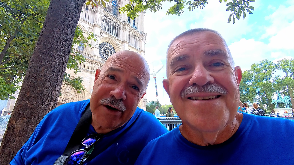 Two smiling older men in blue shirts take a selfie by Notre Dame Cathedral, with trees and tourists behind them.