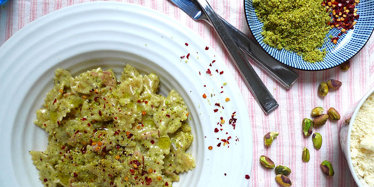 Plate of pasta with pistachio sauce, sprinkled with red chilli flakes. Surrounded by utensils, pistachios, and grated cheese on a striped pink cloth.