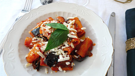 Pasta with tomato sauce, eggplant, and salted ricotta on a white plate. Garnished with basil. Set on a white tablecloth with cutlery.