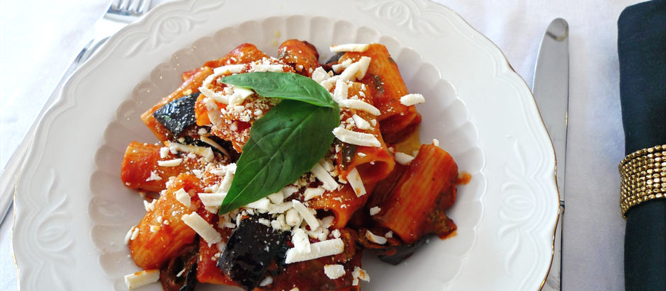 Pasta with tomato sauce, eggplant, and salted ricotta on a white plate. Garnished with basil. Set on a white tablecloth with cutlery.