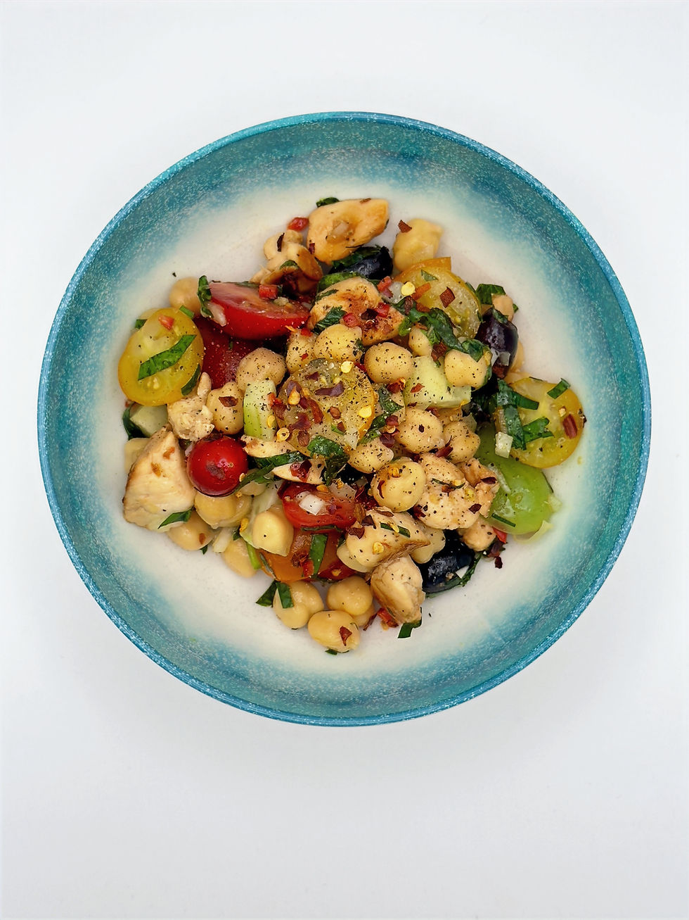 Colorful salad in a blue bowl with tomatoes, chickpeas, cucumbers, and herbs on a white background. Vibrant and fresh appearance.