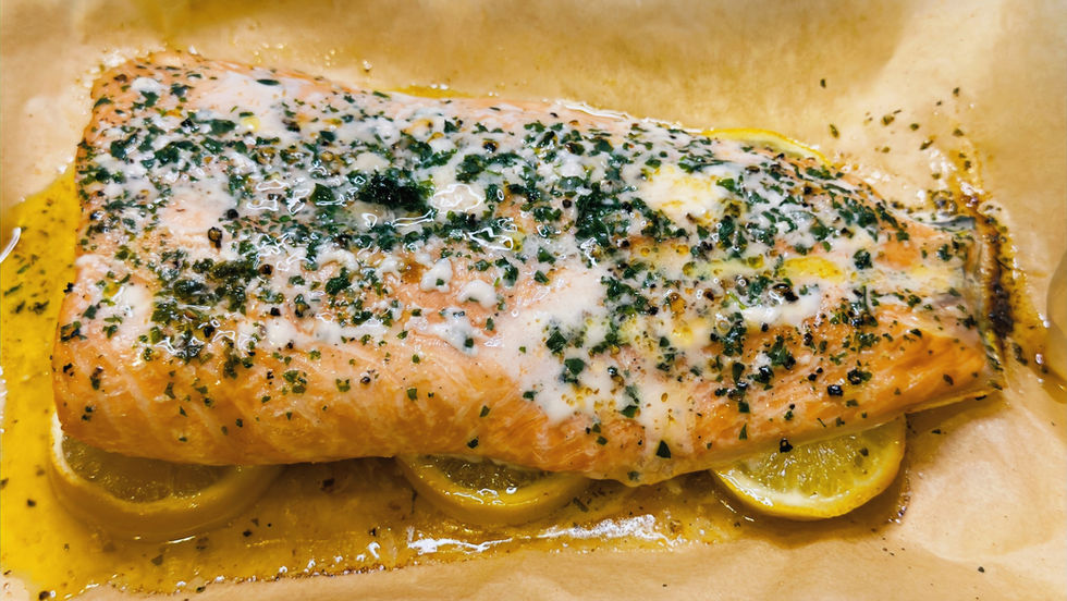 Baked salmon fillet on lemon slices, topped with melted herb butter, on parchment and foil. Background has a speckled countertop.