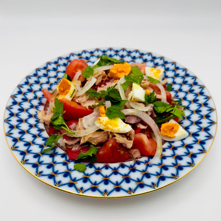 Salad with roasted peppers, tomatoes, eggs, parsley, tuna, and onions on a blue patterned plate with gold accents. White background, colorful and fresh.