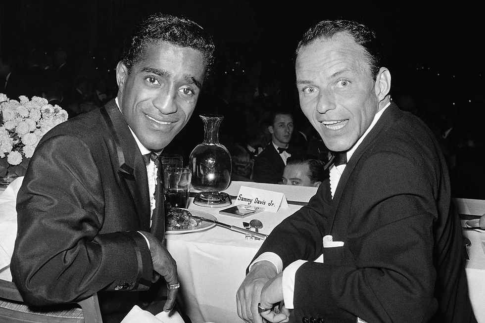Black-and-white photograph of Sammy Davis Jr. and Frank Sinatra seated together at a banquet table, smiling toward the camera.