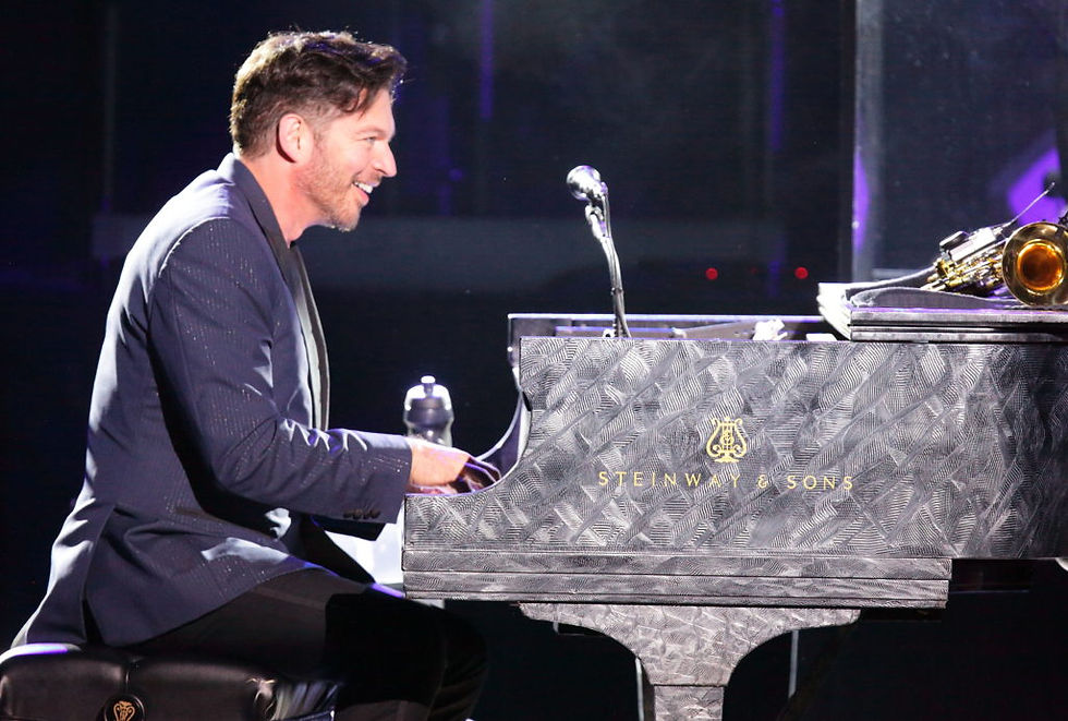 Harry Connick Jr. smiles while playing a Steinway & Sons grand piano during a live concert, with a saxophone resting on top of the piano beside him.