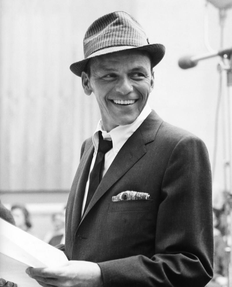 Black-and-white photograph of Frank Sinatra smiling while wearing a fedora, loosened tie, and tailored suit.