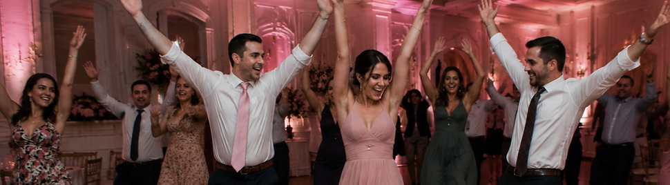 Wedding guests dancing to “YMCA” with arms raised in a ballroom filled with pink uplighting and joyful energy.