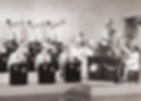 The Count Basie Orchestra performs on stage during the height of the swing era, circa 1940, with Count Basie seated at the piano and the full ensemble arranged behind him.