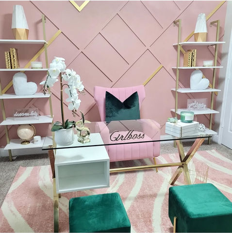 Stylish office interior with a blush pink statement chair, emerald green accents, glass-top desk with gold base, and symmetrical shelving against a pink accent wall with geometric gold inlays.
