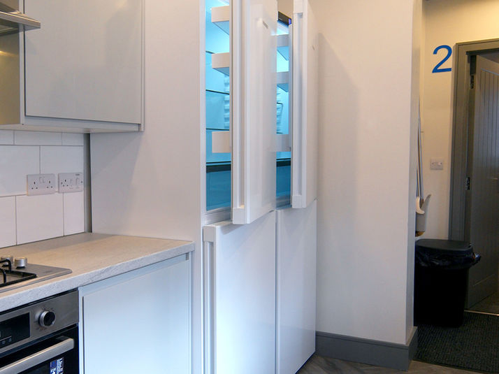 A modern shared kitchen with two tall fridge freezers open to show spacious shelving, set beside sleek white cabinets, a built-in oven and a light grey worktop. A door marked “2” is visible in the hallway beyond.
